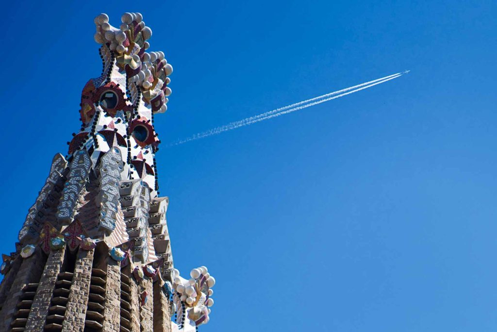 View of Sagrada Familia Tower by Gaudi, and on the background, blue skyes with a jet airplane flying and leaving a track on the sky. 