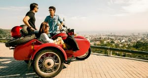 Tour guide explaining Barcelona to a tourist couple while seating on a red sidecar motorcycle by a viewpoint over Barcelona.